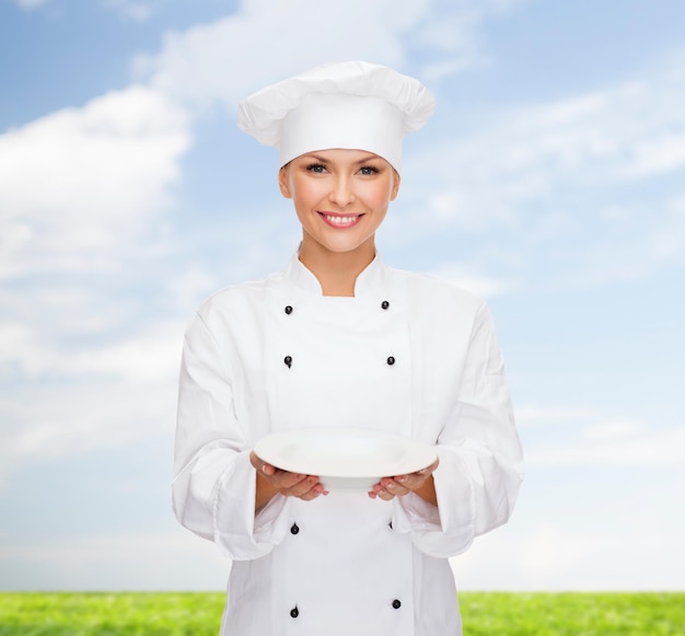 conceito de culinária e comida - chef feminino sorridente, cozinheiro ou padeiro com prato vazio sobre fundo de céu azul e grama