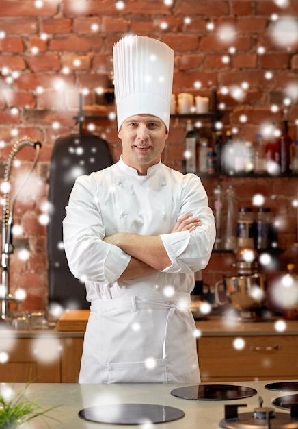 conceito de culinária e pessoas - cozinheiro chef masculino feliz com as mãos cruzadas na cozinha do restaurante sobre o efeito de neve