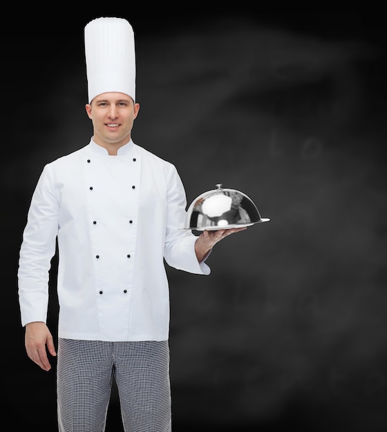 conceito de culinária, profissão e pessoas - cozinheiro chef masculino feliz segurando cloche sobre fundo de quadro de giz preto