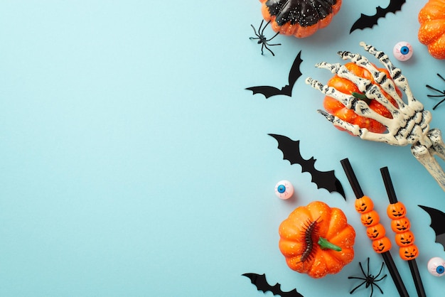 Conceito de decorações de festa de Halloween Foto de vista superior da mão de esqueleto segurando silhuetas de morcego de abóbora aranhas olhos de centopéia e canudos de coquetel em fundo azul pastel isolado com copyspace