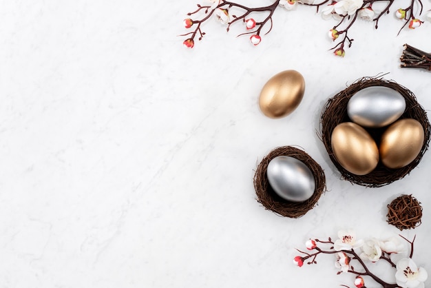 Conceito de design de ovos de Páscoa de ouro e prata no ninho com flor de ameixa branca no fundo da mesa de mármore branco brilhante.