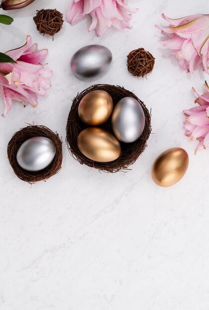 Conceito de design de ovos de páscoa de ouro e prata no ninho com flor de lírio rosa em fundo de mesa de mármore branco brilhante.