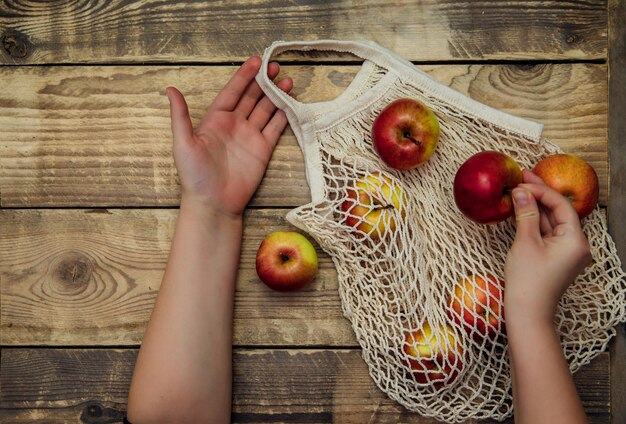 Conceito de desperdício zero, sacolas de compras de malha reutilizáveis. sacola ecológica feita de tecido de algodão natural em um fundo de madeira. a mão de uma mulher segura uma maçã madura.