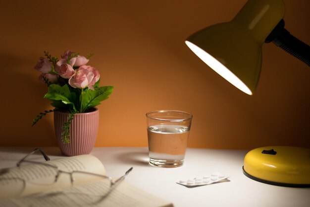 Conceito de detalhe da mesa de cabeceira com copos de livro vaso de flores de livro copo de remédios de lâmpada de água