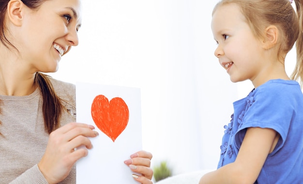 Conceito de dia das mães. Filha criança parabeniza a mãe e dá seu cartão postal com formato de coração vermelho. Mãe e menina felizes sorrindo e abraçando. Diversão em família e férias.