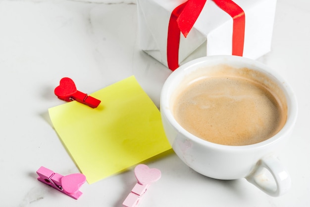 Conceito de dia dos namorados, caixa de presente com fita vermelha, caneca de café, nota de papel em branco para parabéns com pinos em forma de coração, branco, copyspace vista superior