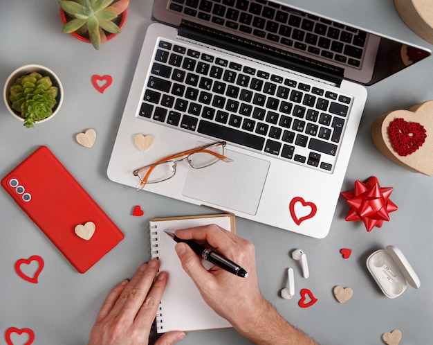 Conceito de dia dos namorados com gadgets modernos e mãos escrevendo na vista superior de um caderno na mesa cinza