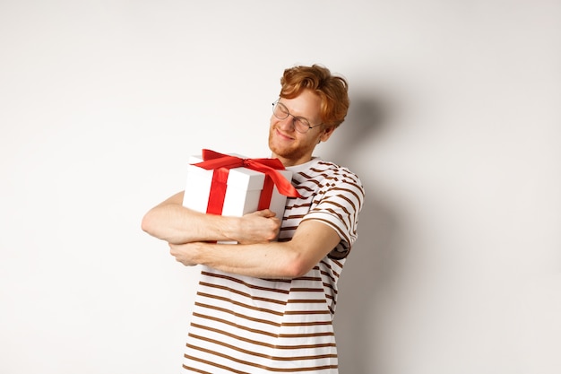 Conceito de dia dos namorados e feriados. Homem ruivo feliz abraçando sua caixa de presente e sorrindo encantado, de pé sobre um fundo branco