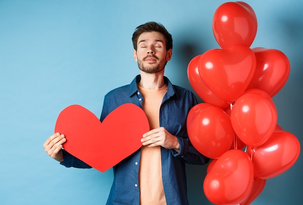 Conceito de dia dos namorados. homem sonhando com o amor verdadeiro, segurando o recorte de coração vermelho e em pé perto de balões românticos, fundo azul.