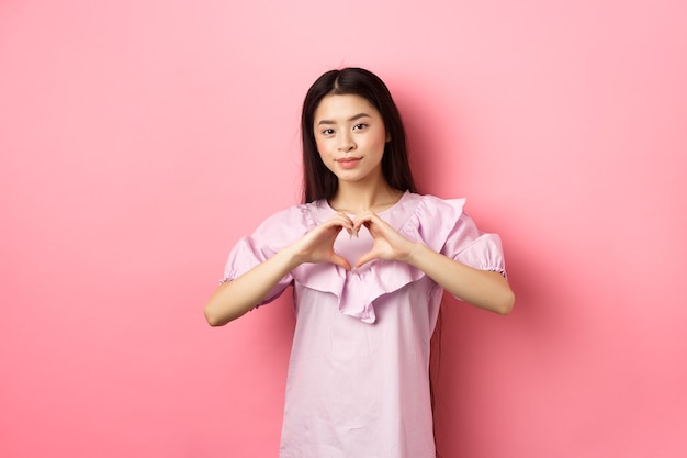 Conceito de dia dos namorados. Linda menina adolescente japonesa mostra gesto de coração e sorriso bonito, expressa amor e simpatia ao amante, em um vestido romântico em fundo rosa.