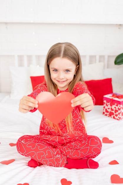 Conceito de dia dos namorados uma linda menina criança está sentada na cama em casa de pijama vermelho e segurando seu coração nas mãos e sorrindo parabenizando pelo feriado