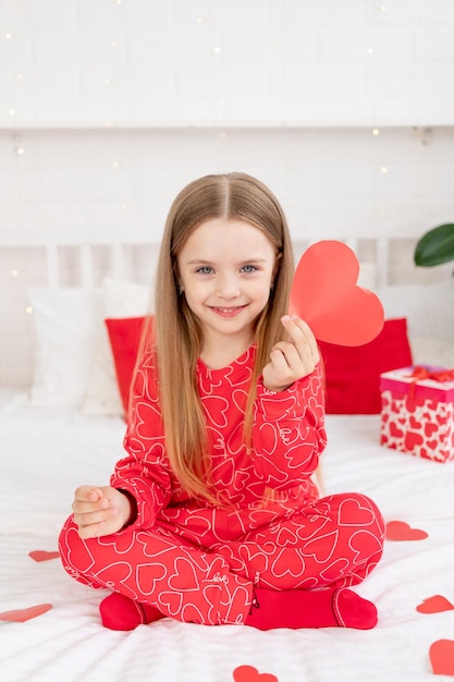 Conceito de dia dos namorados uma linda menina criança está sentada na cama em casa de pijama vermelho e segurando seu coração nas mãos e sorrindo parabenizando pelo feriado