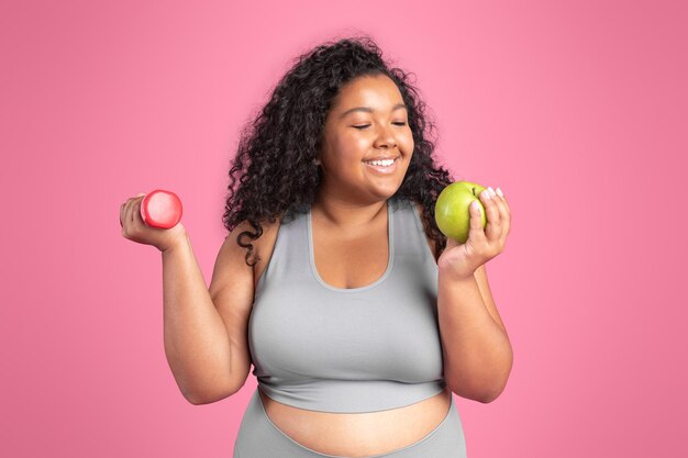 Conceito de dieta saudável feliz corpo negro mulher positiva segurando halteres e maçã desfrutando de frutas frescas