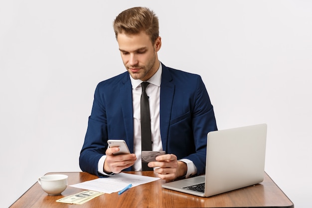 Conceito de dinheiro, negócios e finanças. bonito jovem empresário masculino sentado na mesa do escritório usando o smartphone para pagar as contas, fazer compras on-line, segurando o cartão de crédito, fundo branco