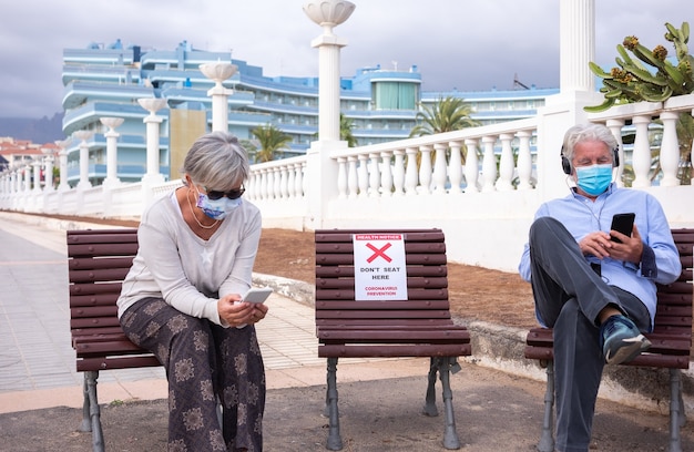 Conceito de distanciamento social. Duas pessoas seniores sentadas ao ar livre em um banco permitido, usando telefone celular