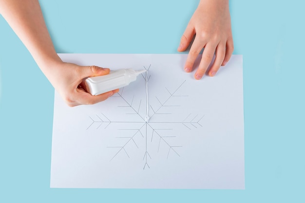 Conceito de diy e criatividade infantil. instruções passo a passo: como fazer desenho de floco de neve com cola e sal. passo 3 as mãos da criança aplicam cola no desenho.