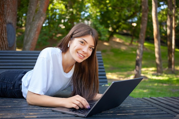 Conceito de educação e tecnologia de trabalho remoto jovem estudante bonita sorridente deitada no banco no parque usando fones de ouvido sem fio, anotando e fazendo agendamento no laptop copie o espaço