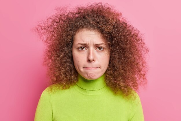 Conceito de emoções e reações humanas de pessoas. Preocupada insatisfeita com cabelos encaracolados jovem europeia quer chorar pressiona os lábios vestidos com gola olímpica verde casual isolada sobre a parede rosa.