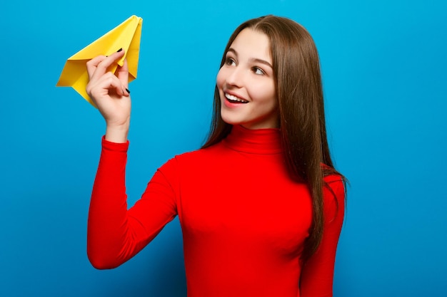 Conceito de emoções, pessoas, beleza, moda e estilo de vida, viagens, turismo e pessoas - retrato de uma linda mulher segurando um avião de papel amarelo. Sobre um fundo azul.