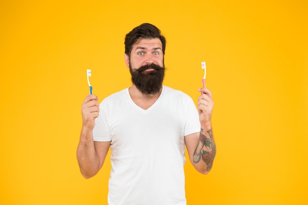 Conceito de escovar os dentes Higiene dos dentes Tecnologia de limpeza ultra-sônica dos dentes Homem barbudo segura escovas de dentes fundo amarelo Cara segura duas escovas de dentes Hipster rosto sorridente feliz cuida da higiene