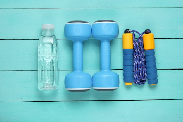 Conceito de esporte de estilo liso leigo. Halteres, pular corda, garrafa de água. Equipamento desportivo em fundo azul de madeira.