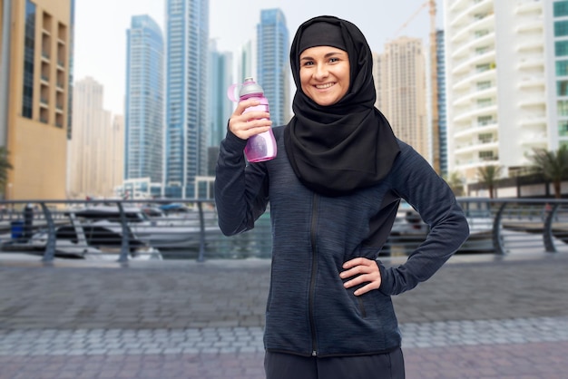 conceito de esporte, fitness e pessoas - mulher muçulmana sorridente feliz em hijab com garrafa de água sobre o fundo da rua da cidade de dubai