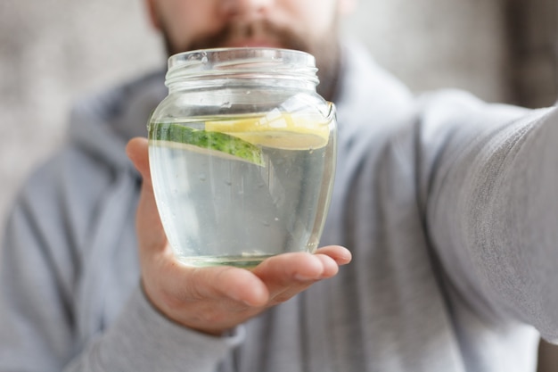 Conceito de esporte, saúde, pessoas, emoções, 4k e estilo de vida - foto portátil de um homem encapuzado bebendo água, em câmera lenta. bebida fresca com fatias de limão e pepino no copo