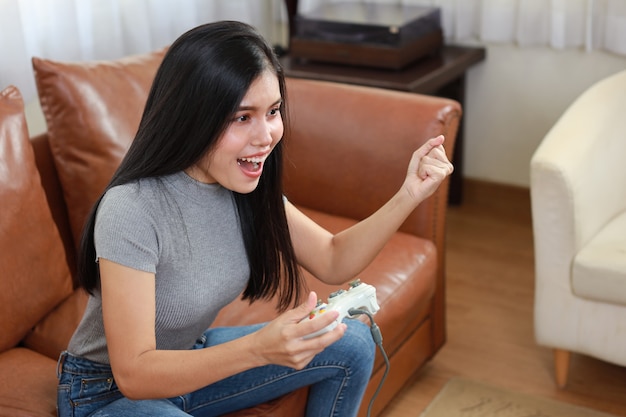 Conceito de estação de console de jogos vdo. mulher asiática ativa sentada no sofá, segurando o joystick e jogando um jogo emocionante. linda garota parecia animada com o console do controle do jogo