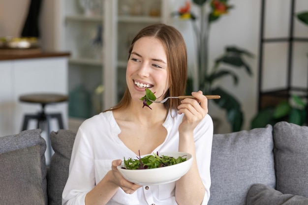 Conceito de estilo de vida de comida saudável. Jovem e linda mulher comendo salada verde fresca, sentada em casa no sofá