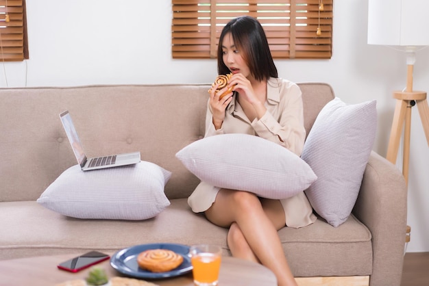 Conceito de estilo de vida de relaxamento Jovem asiática assistindo filme no laptop e comendo pão no sofá