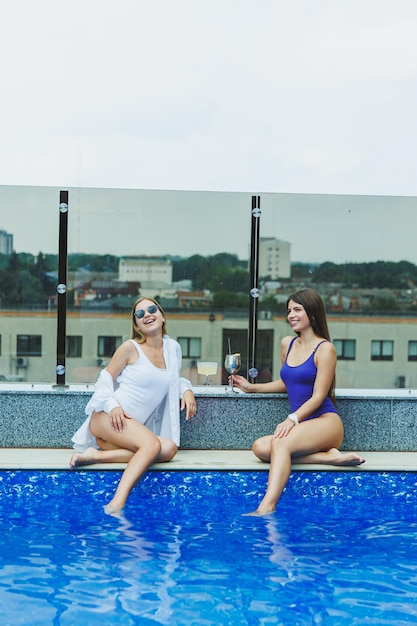 Conceito de estilo de vida Duas mulheres atraentes em óculos de sol bebendo coquetéis à beira da piscina Vista lateral de glamourosas senhoras quentes em trajes de banho se divertindo na piscina turquesa