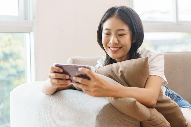 Conceito de estilo de vida em casa jovem mulher sentada no sofá e abraçando o travesseiro para jogar no smartphone em casa