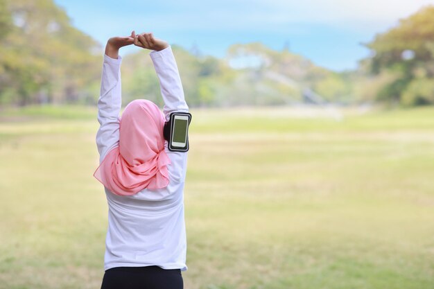 Foto conceito de estilo de vida exercício público, música fitness mulher de fones de ouvido sem fio e telefone celular. vista traseira atlética jovem muçulmana asiática no sportswear em pé, estendendo-se após treino