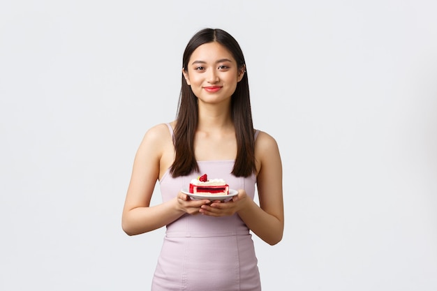 Conceito de estilo de vida, feriados, celebração e comida. Linda garota asiática em um vestido de noite, desfrutando de comer um bolo doce e delicioso