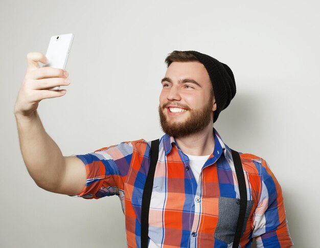 Conceito de estilo de vida um jovem com barba na camisa segurando o celular e fazendo foto de si mesmo em pé contra um fundo cinza