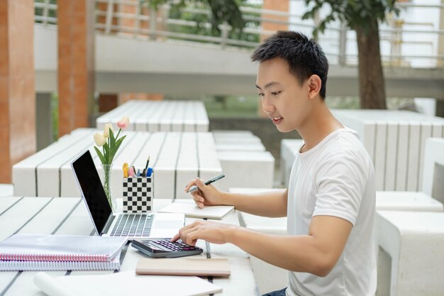 Conceito de estudo online um estudante do sexo masculino em uma camiseta branca estudando online usando seu novo laptop branco e a calculadora na aula de contabilidade.