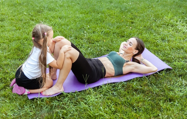 Foto conceito de família ativa mãe em forma treina os músculos abdominais enquanto está deitada no tapete com sua filha pequena ao ar livre no parque estilo de vida saudável