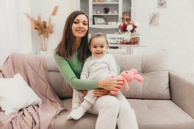 Conceito de família, bebê e maternidade - jovem mãe feliz e sorridente com o bebê no sofá em casa. Dia das Mães