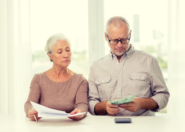 Conceito de família, economia, idade e pessoas - casal sênior com papéis, dinheiro e calculadora em casa