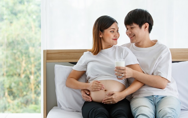 Conceito de família feliz e saudável. Foto de um jovem casal sentado numa cama juntos. Um jovem marido sorrindo e segurando um copo de leite e dando com amor a uma esposa grávida.