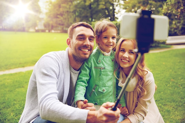 conceito de família, infância, tecnologia e pessoas - feliz pai, mãe e filha tirando foto pelo smartphone selfie stick no parque