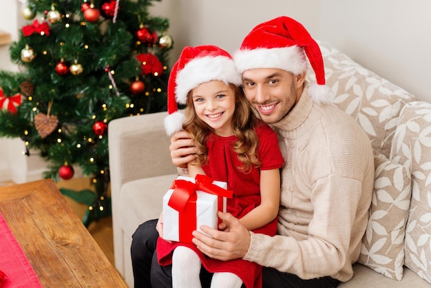 Conceito de família, natal, natal, inverno, felicidade e pessoas - pai e filha sorrindo com chapéus de ajudante de papai noel segurando uma caixa de presente