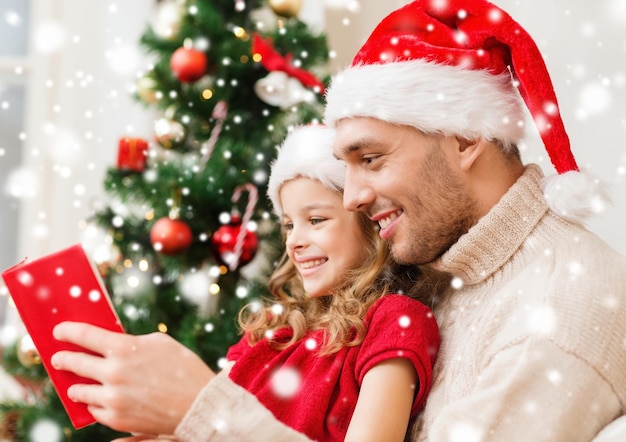 conceito de família, natal, pessoas e feriados - sorrindo pai e menina em chapéus de Papai Noel lendo livro ou bíblia em casa