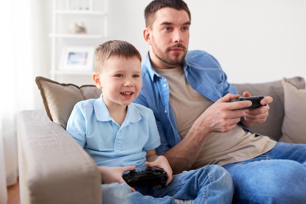 conceito de família, paternidade e pessoas - pai feliz e filho pequeno com gamepads jogando videogame em casa