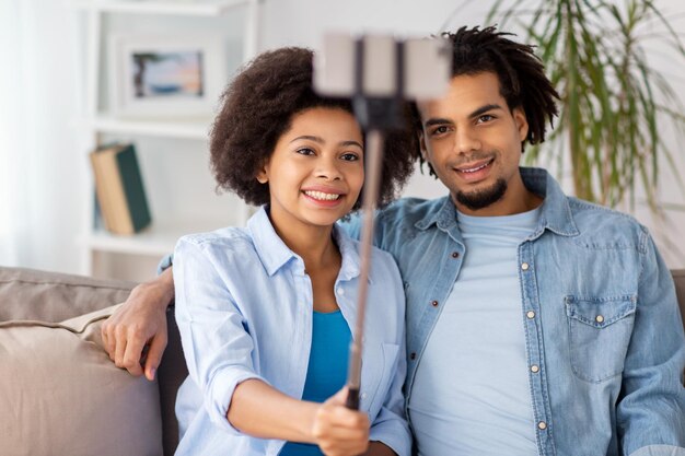 conceito de família, tecnologia e pessoas - casal feliz com smartphone e bastão de selfie tirando fotos em casa