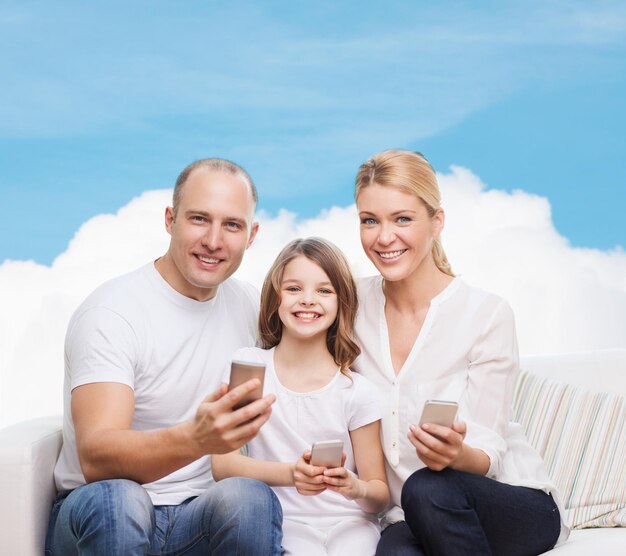 conceito de família, tecnologia e pessoas - sorrindo mãe, pai e filha com smartphones sobre céu azul e fundo de nuvem branca