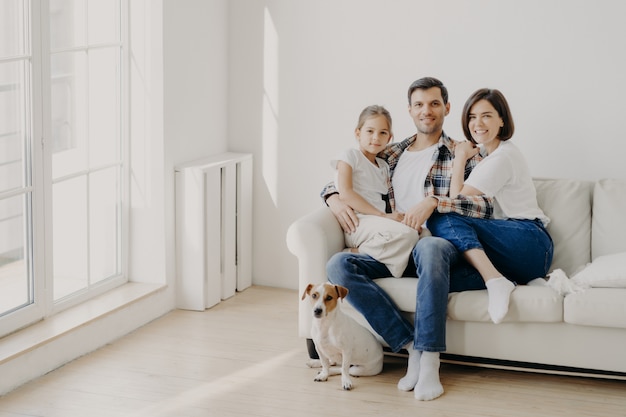 Conceito de família, união e relacionamento. O homem feliz abraça a filha e a esposa, senta-se no sofá branco confortável na sala vazia, o animal de estimação senta-se no chão, faz o retrato de família para uma memória longa