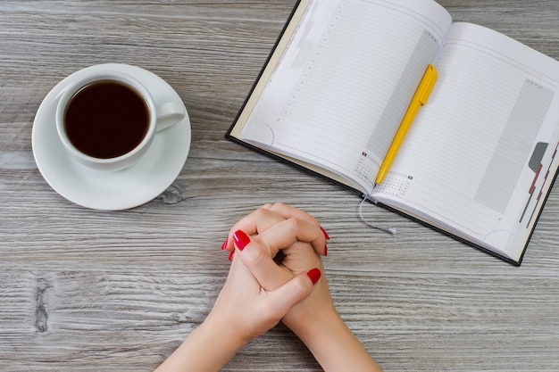 Conceito de fazer uma escolha entre educar e descansar. foto da vista superior das mãos cruzadas de uma mulher, xícara de café, bloco de notas aberto com caneta na mesa de madeira