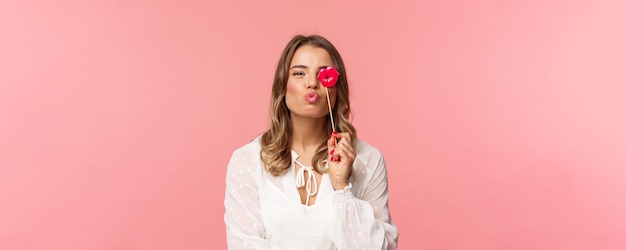 Conceito de felicidade e celebração da primavera Closeup retrato de uma linda garota loira feminina no vestido branco, dobrando os lábios no beijo e segurando o bastão de papelão festejando se divertindo no fundo rosa