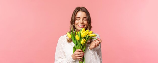 Conceito de felicidade e celebração da primavera retrato aproximado de uma linda garota loira romântica cheirando o cheiro de lindas tulipas amarelas fecha os olhos e sorrindo feliz fundo rosa em pé